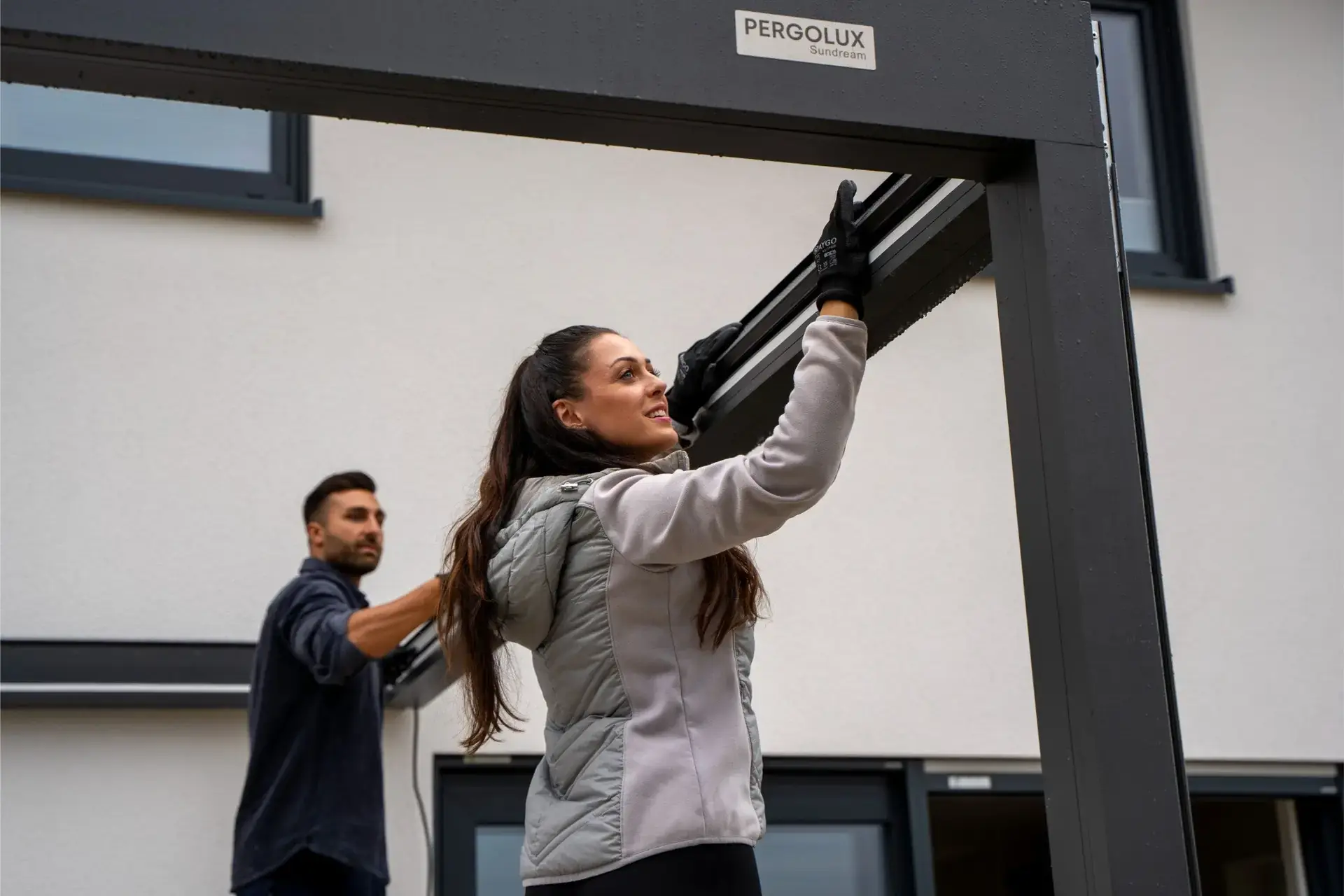 Two people fitting the beam of a dark gray louvered pergola kit against the wall of a white house