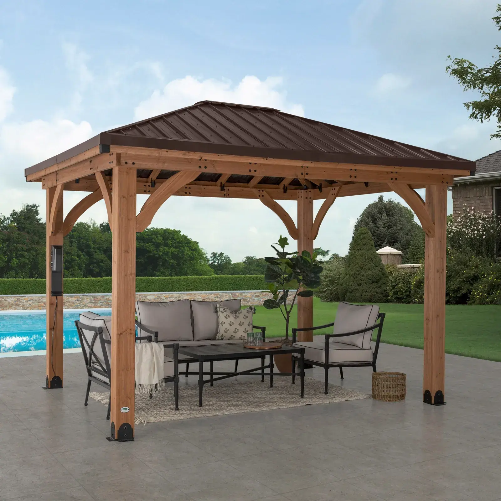Backyard Discovery Barrington cedar gazebo with a brown steel hip roof on thick cedar posts, set against trees and a hedge
