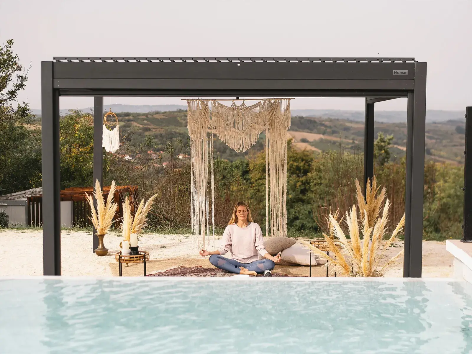 Hansø Pro+ pergola with a dark gray powder-coated aluminum frame over a lounge seating area, louvers open, hillside beyond