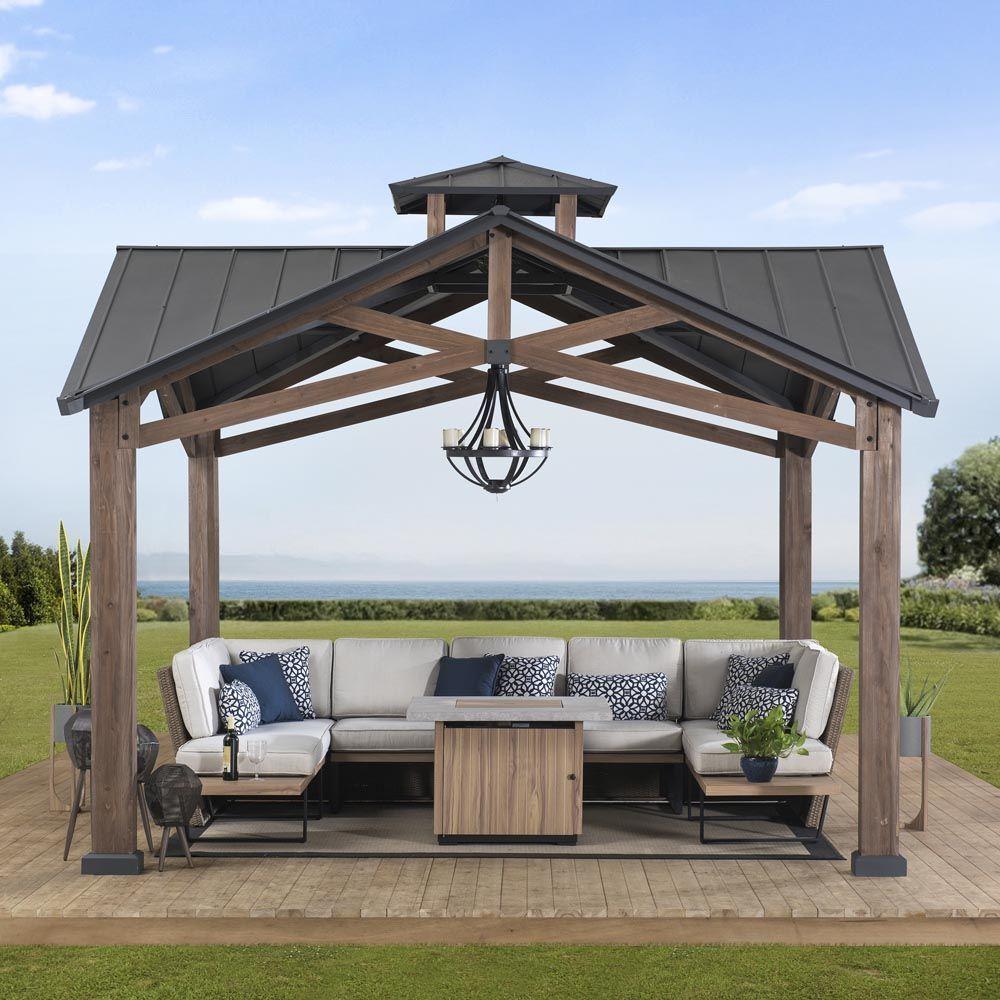 Sunjoy Bella 12.5x12.5 gazebo on decking above a shoreline, sheltering a corner sofa and fire table with a lantern on the ceiling hook