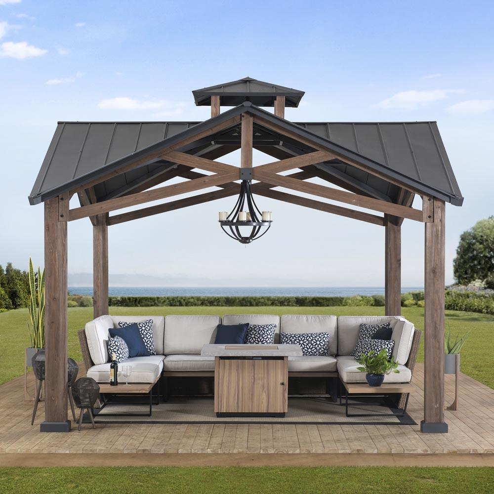 Sunjoy Bella 12.5x12.5 gazebo on decking above a shoreline, sheltering a corner sofa and fire table with a lantern on the ceiling hook