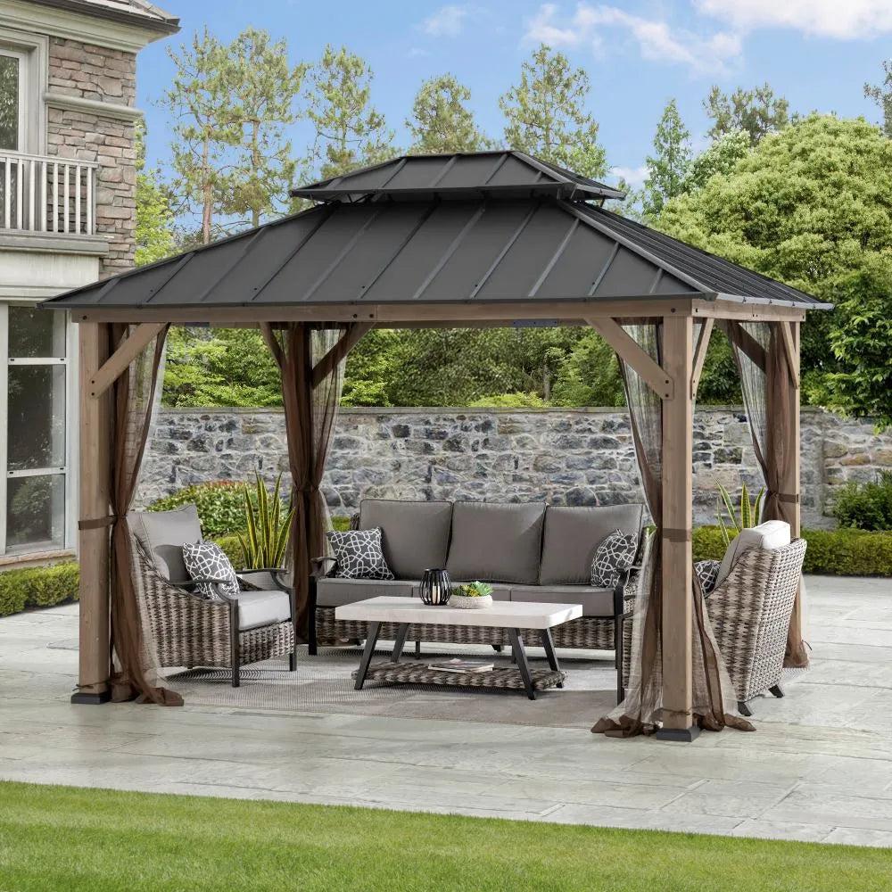 Sunjoy Cedar Dual-Track 10x12 gazebo on a paved terrace beside a stone wall, sheltering a sofa and coffee table with the netting drawn back