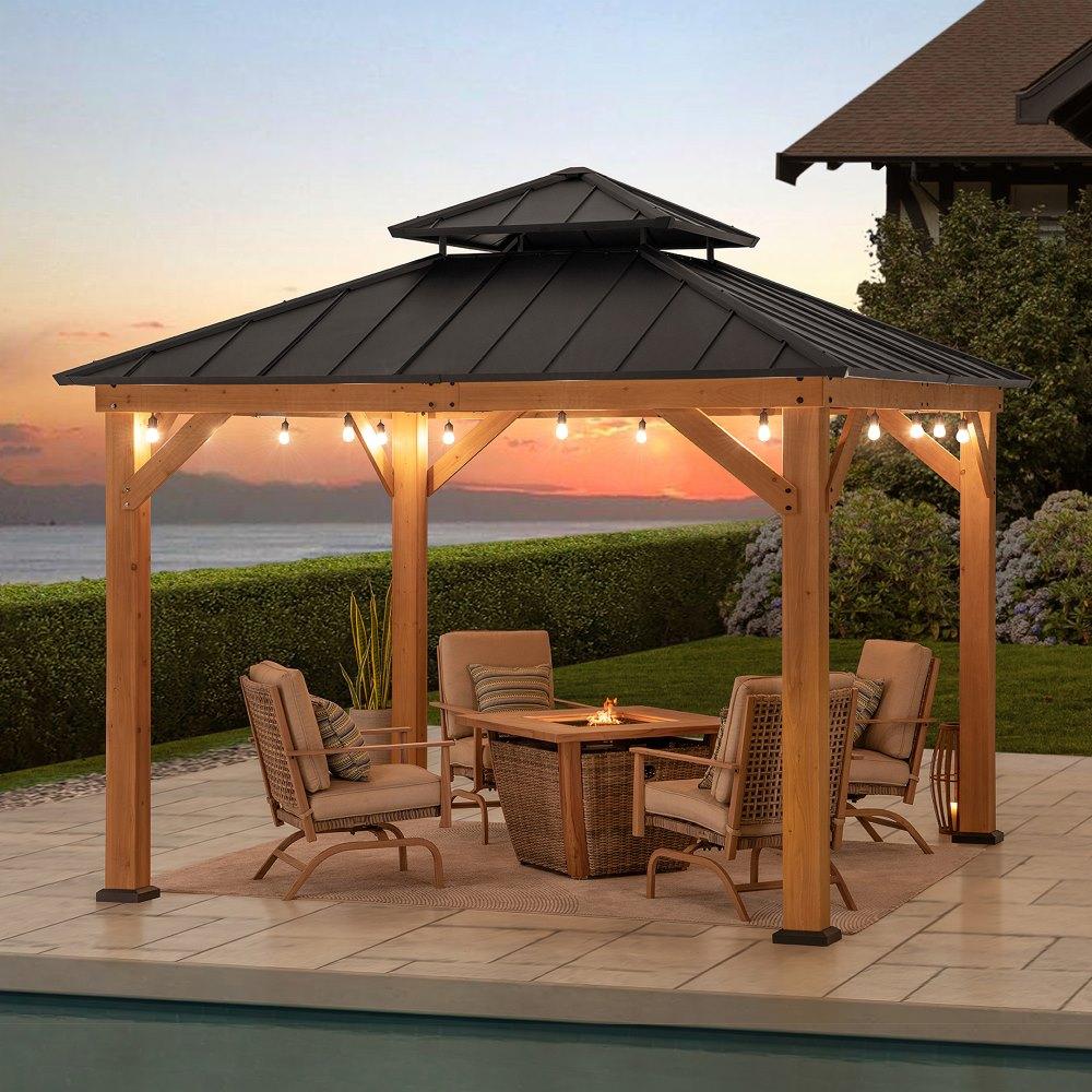 Sunjoy Classic Wooden Hardtop 11x11 gazebo at sunset over a dining table and fire table, string lights along the beams