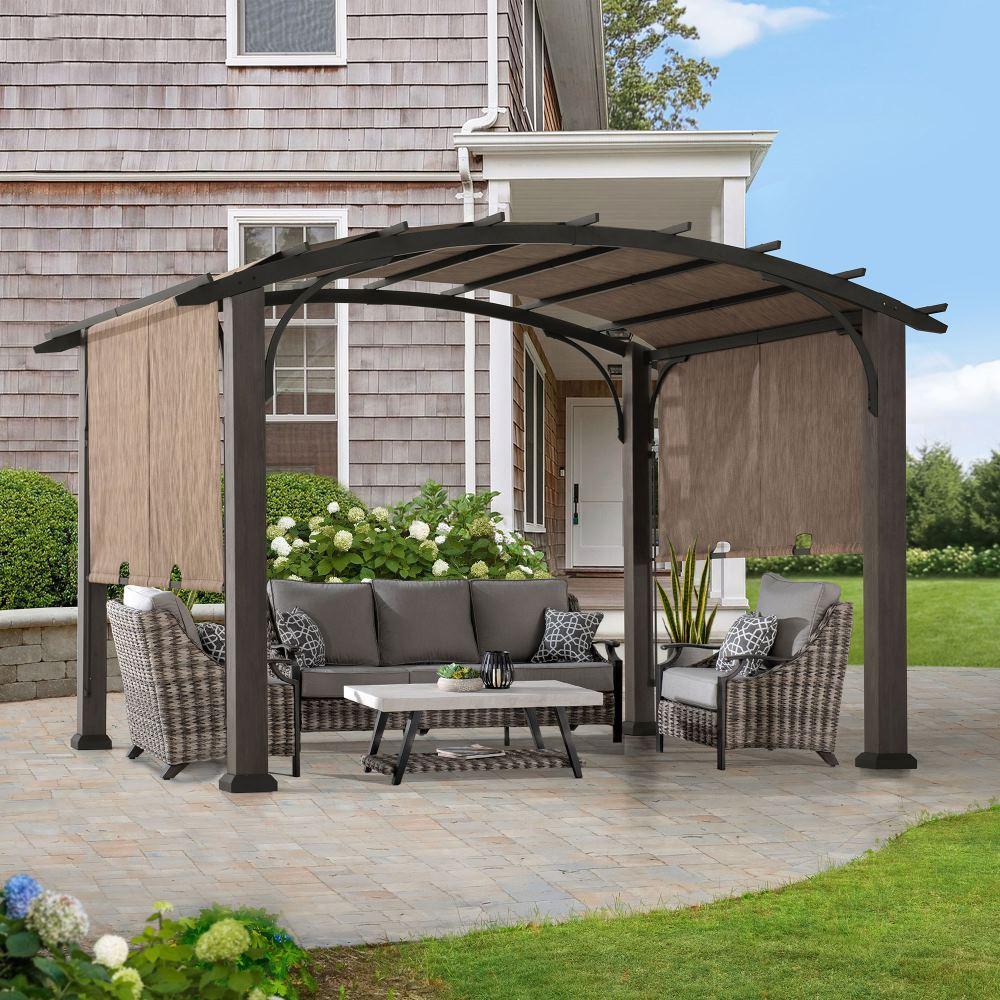 Sunjoy curved-roof retractable pergola on a paver patio beside a shingled house, shading a grey wicker sofa set