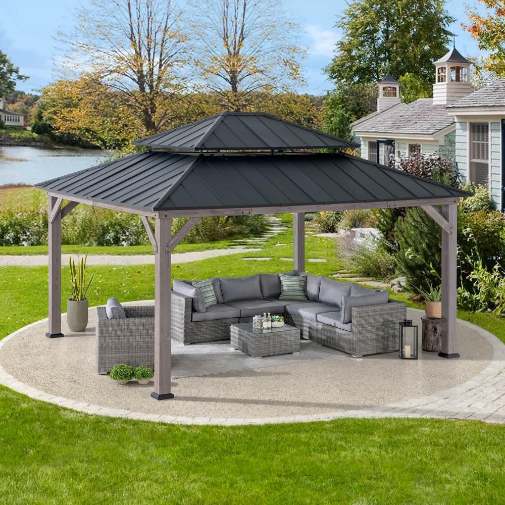 Sunjoy Grand Double-Tier 13x15 gazebo on gravel beside a lake, sheltering a large rattan corner sofa
