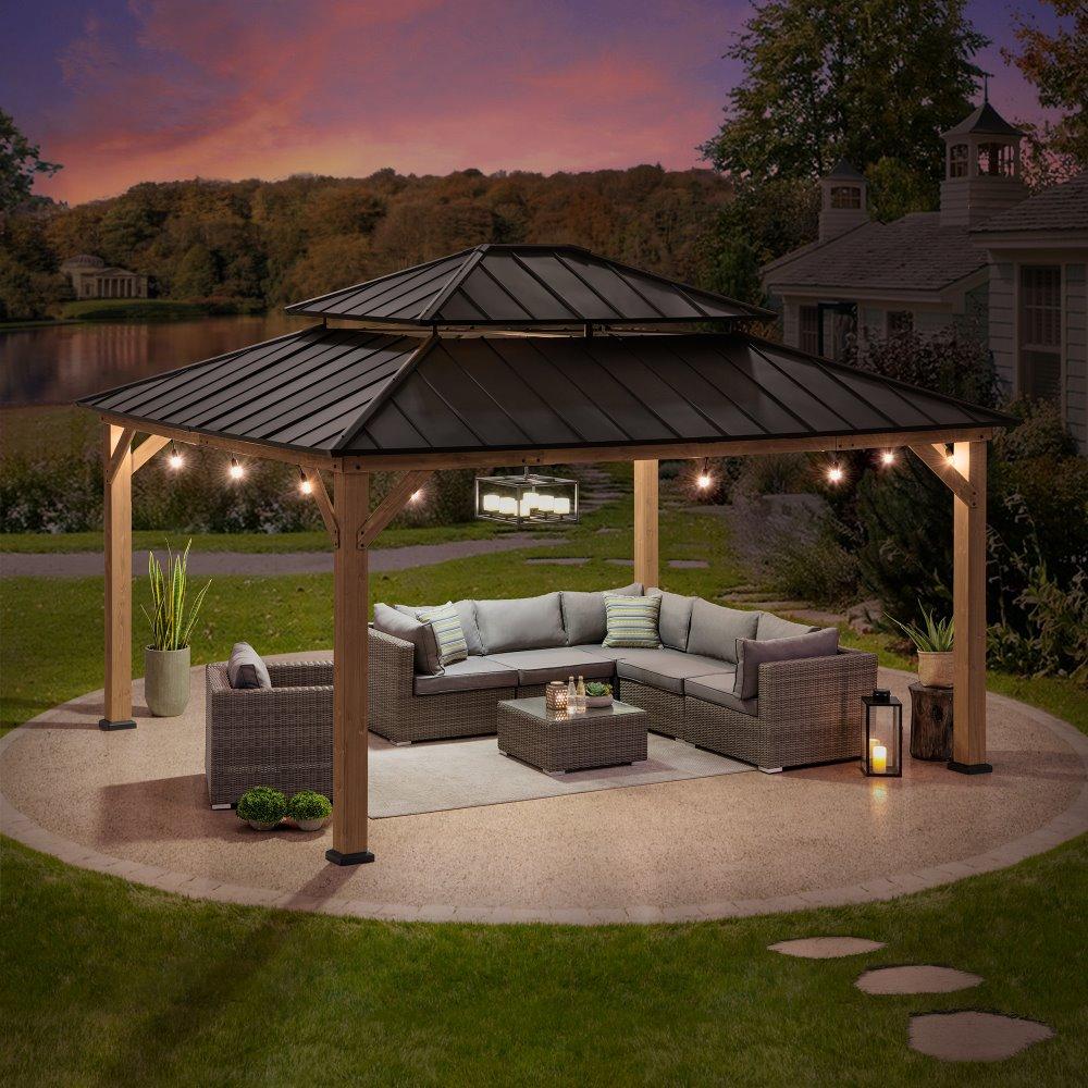 Sunjoy Grand Double-Tier 13x15 gazebo in weathered cedar lit at dusk, string lights along the beams above a corner sofa