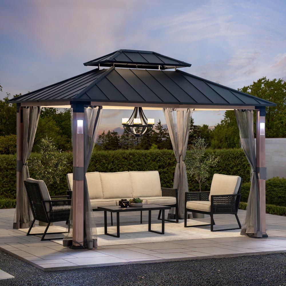 Sunjoy Kirkfield 10x12 metal gazebo on a paved patio sheltering a sofa and armchairs, with a pendant lantern hung from the ceiling hook