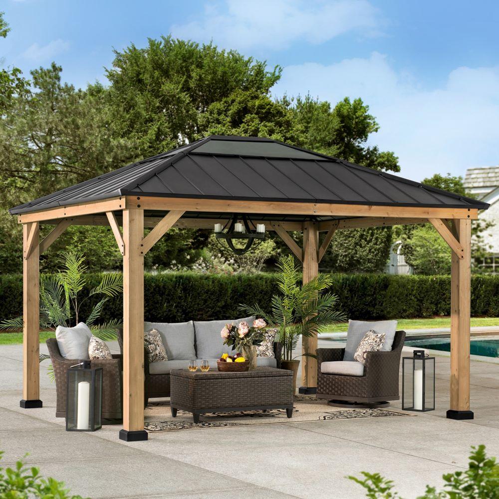 Sunjoy 11x13 wooden skylight gazebo on a paved patio beside a pool, sheltering a corner sofa and fire table