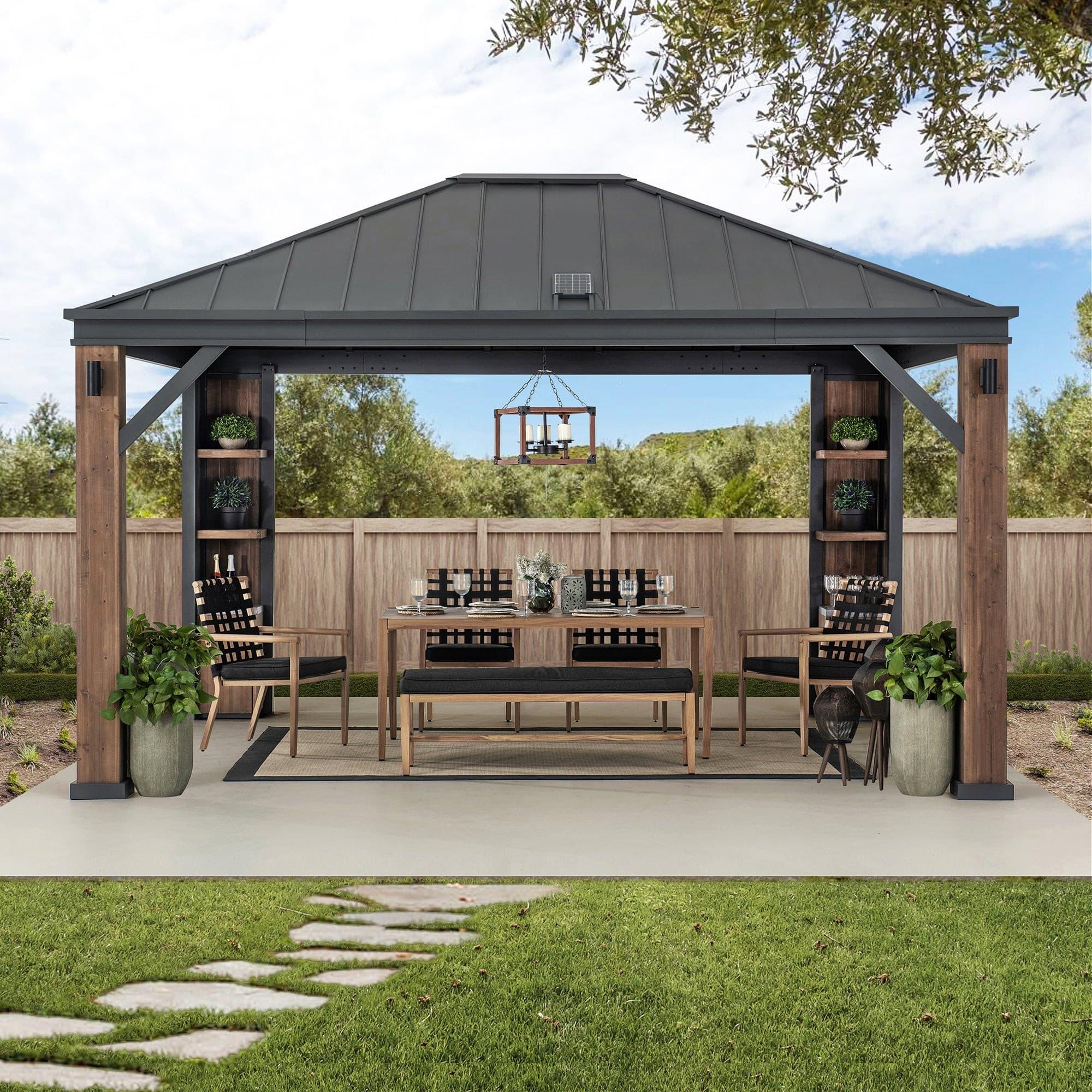 Sunjoy Solaria Smart Tech 12x14 gazebo on a lawn beside a fence, sheltering a long dining table with the corner cabinets stocked
