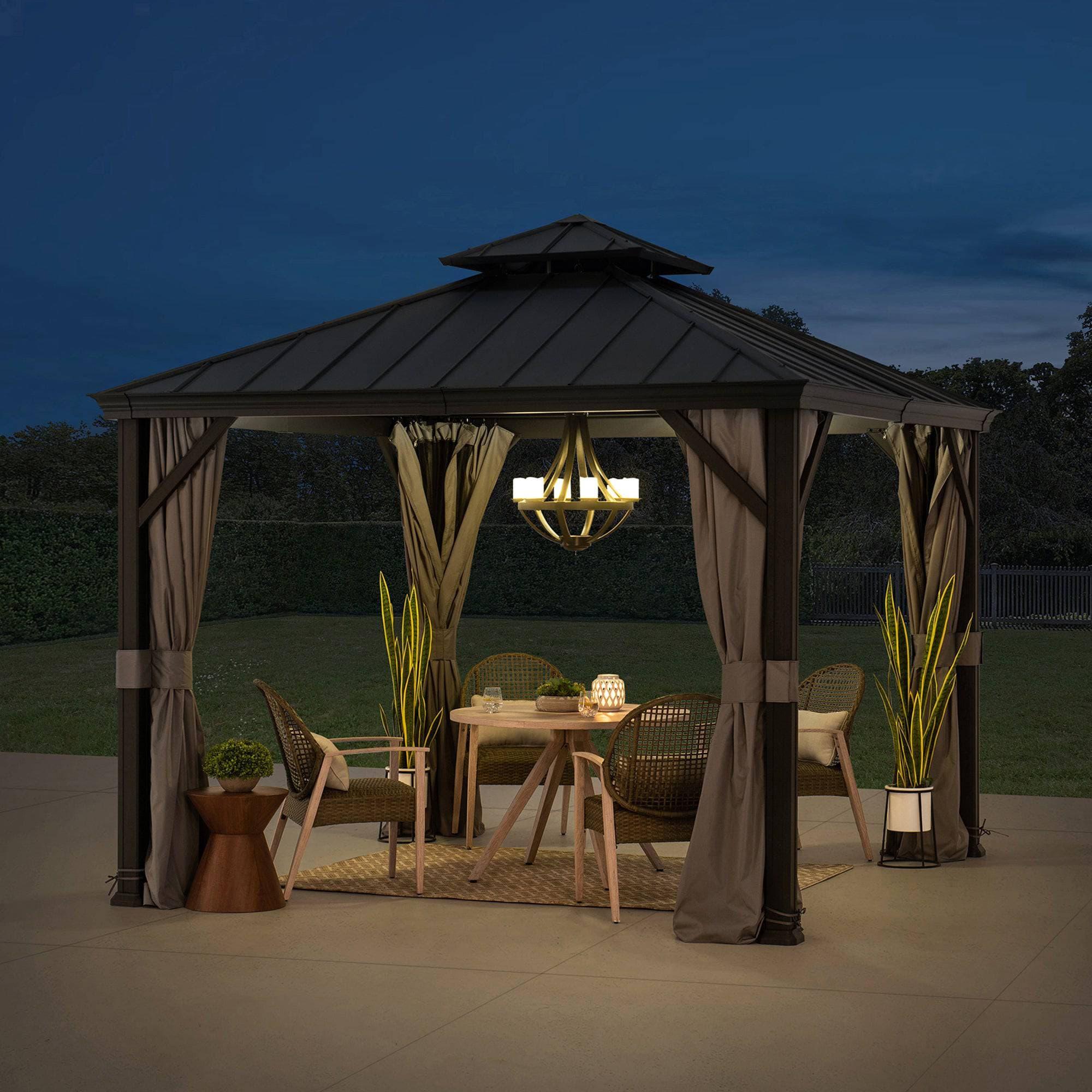 Sunjoy Summit Minimalist 10x10 aluminum gazebo lit at night over a dining table, with a pendant lantern hung from the ceiling hook