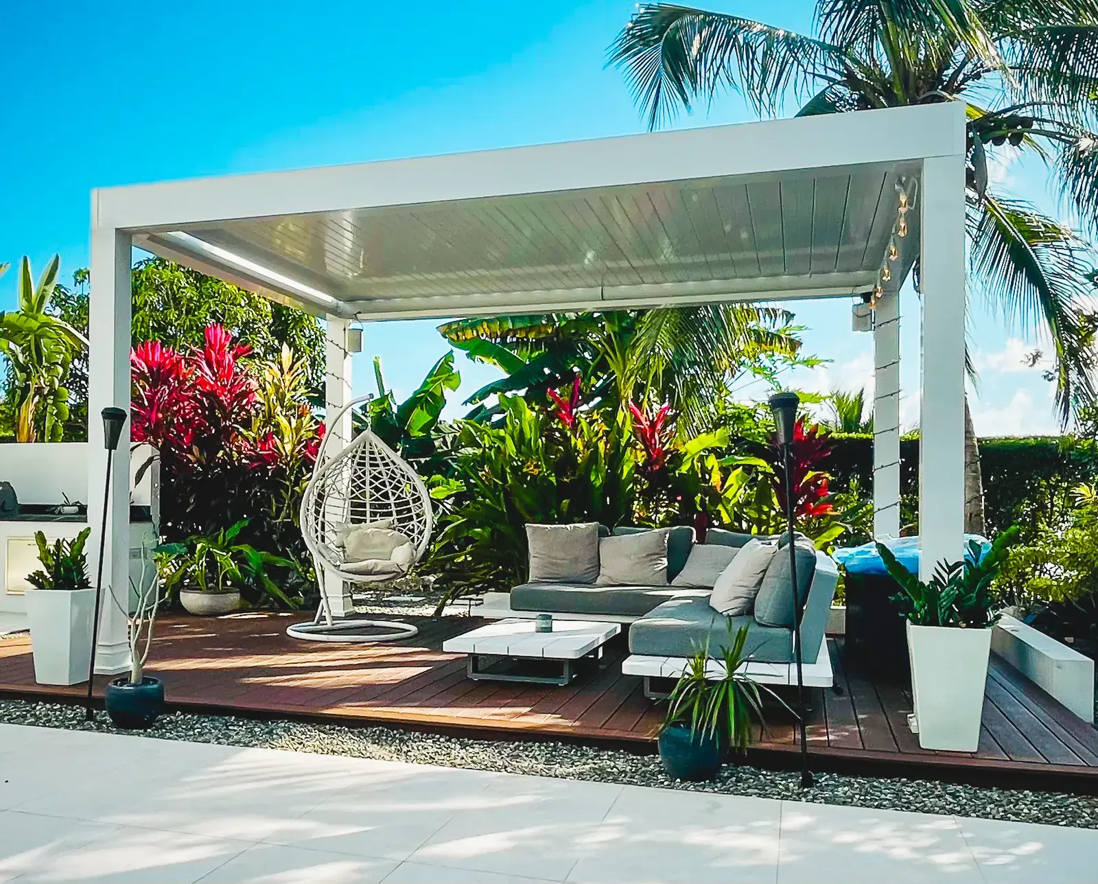 The Luxury Pergola in white with louvers partly open, over a tropical deck with a hanging chair and outdoor sofa