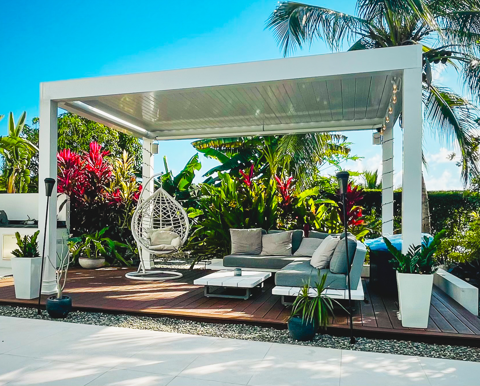 The Luxury Pergola in white with louvers partly open, over a tropical deck with a hanging chair and outdoor sofa