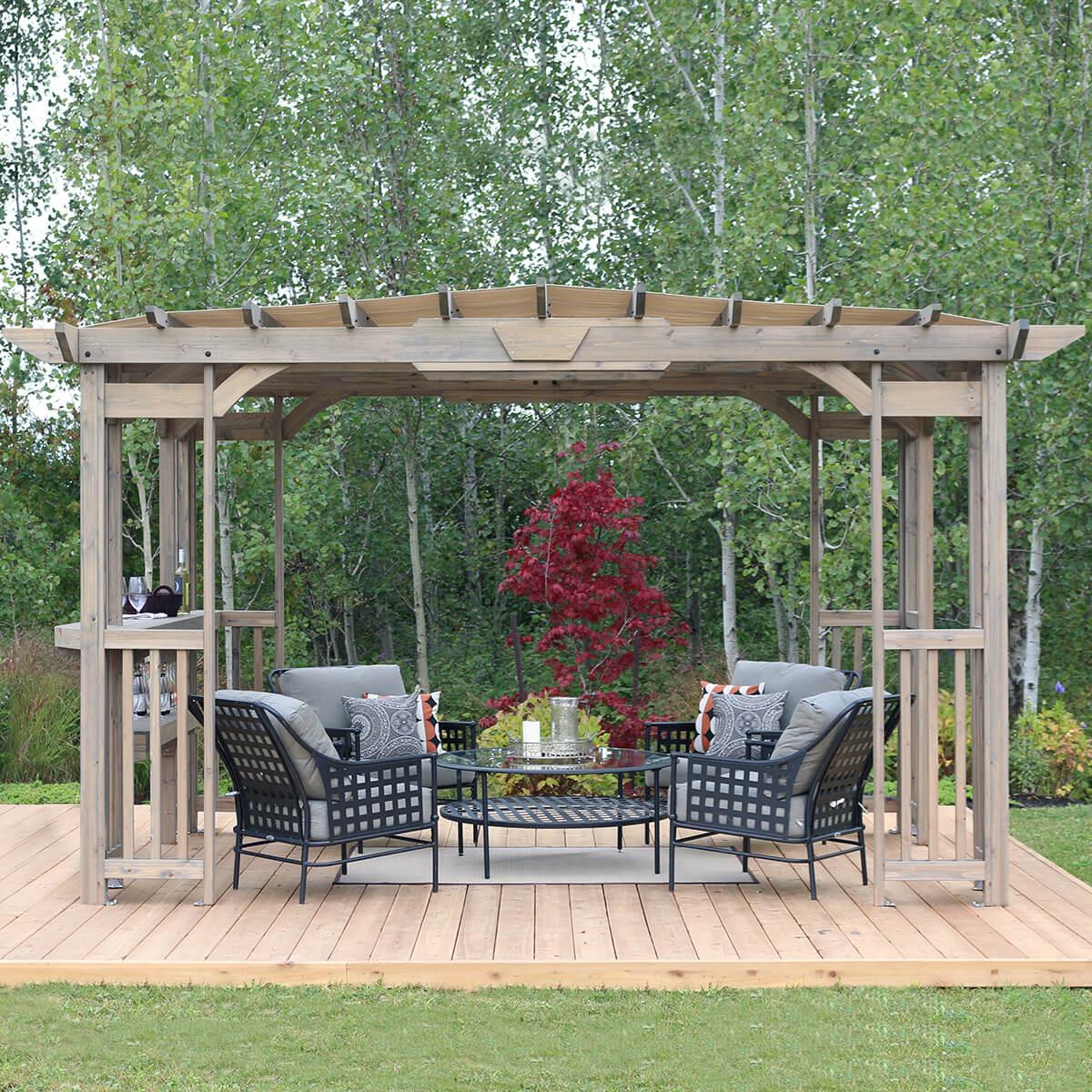 Yardistry Madison Pergola in Timber Grey cedar on a wooden deck, sheltering a patio lounge set in front of a wooded garden