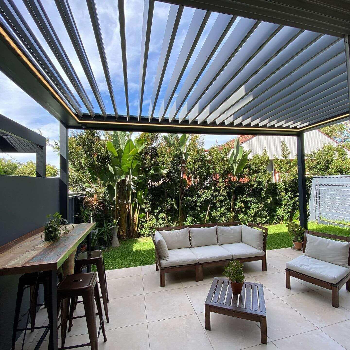 Louvers tilted open on a motorized pergola, with a strip of LED lighting along the beam, over an outdoor sofa in a planted garden